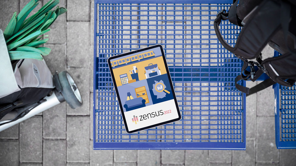 Dieses Bild zeigt einen Einkaufswagen mit einem Tablet auf dem das Zensuslogo 2022 zu erkennen ist, einen Rucksack sowie einen Einkaufstrolley.