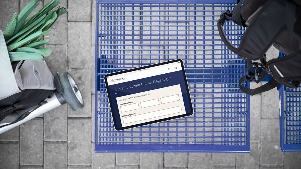 Dieses Bild zeigt einen Einkaufswagen mit einem Tablet auf dem die Anmeldung zum Fragebogen des Zensus 2022 zu erkennen ist, einen Rucksack sowie einen Einkaufstrolley.