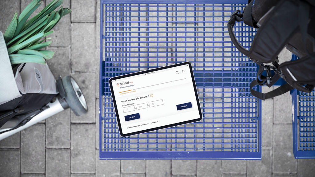 Dieses Bild zeigt einen Einkaufswagen mit einem Tablet auf dem ein Ausschnitt des Fragebogens zum Zensus 2022 zu erkennen ist, einen Rucksack sowie einen Einkaufstrolley.